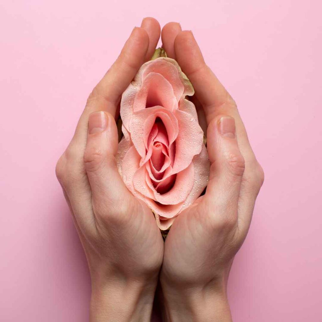 yoni massage symboliseret ved hænder, der omslutter en lyserød blomst på en rosa baggrund, med fokus på feminin omsorg og selvkærlighed.