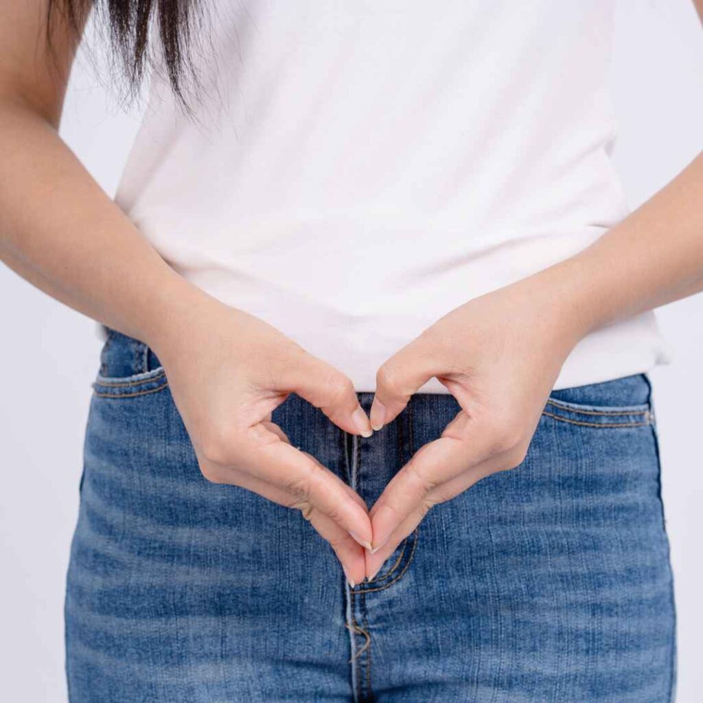 kvinde iført jeans og hvid top former et hjerte med hænderne foran underlivet som symbol på kærlighed og omsorg for kvinders intime sundhed.