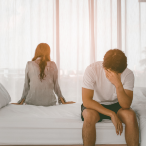 manglende sexlyst illustreret ved et par, der sidder adskilt på sengen med følelsesmæssig afstand, hvor manden ser frustreret ud og kvinden vender ryggen til.