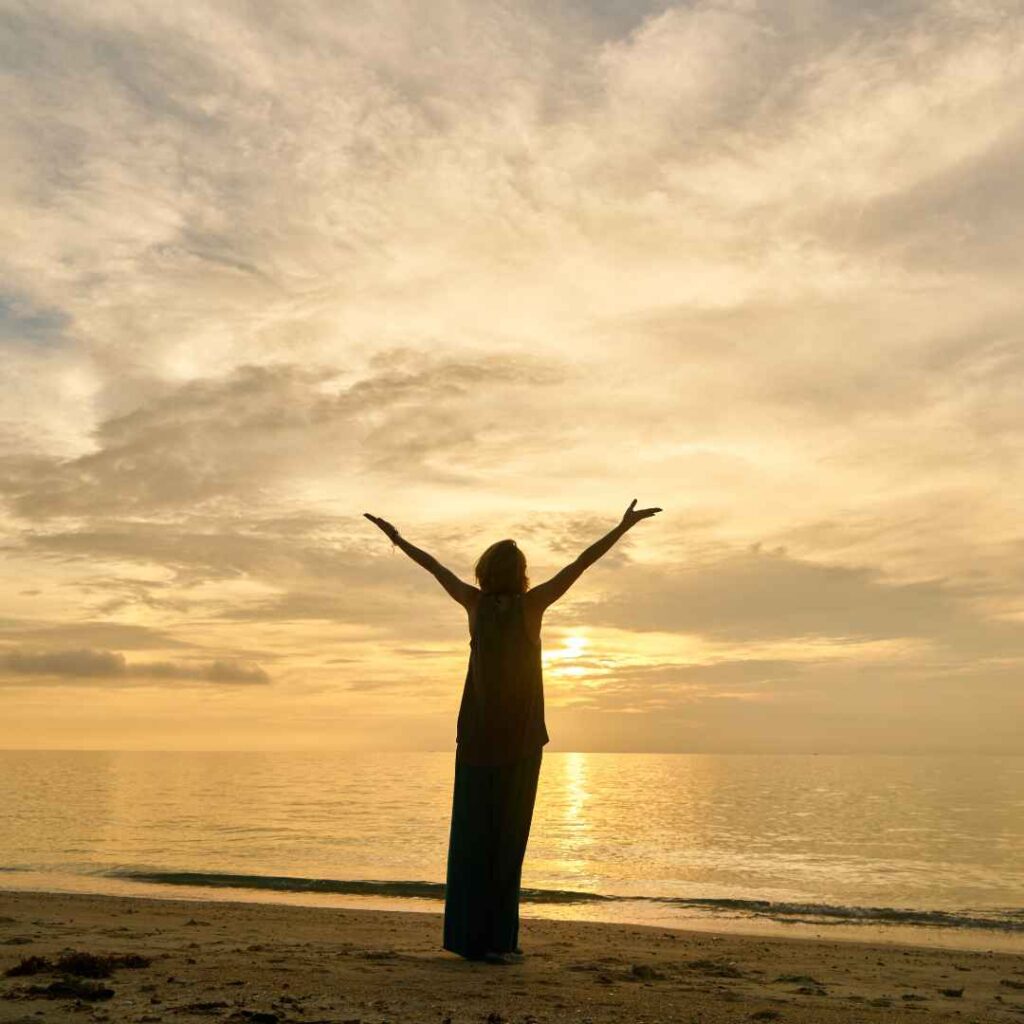 kvinde står på stranden ved solnedgang med arme løftet mod himlen, symboliserer frihed og forbindelse til naturen og kroppen.
