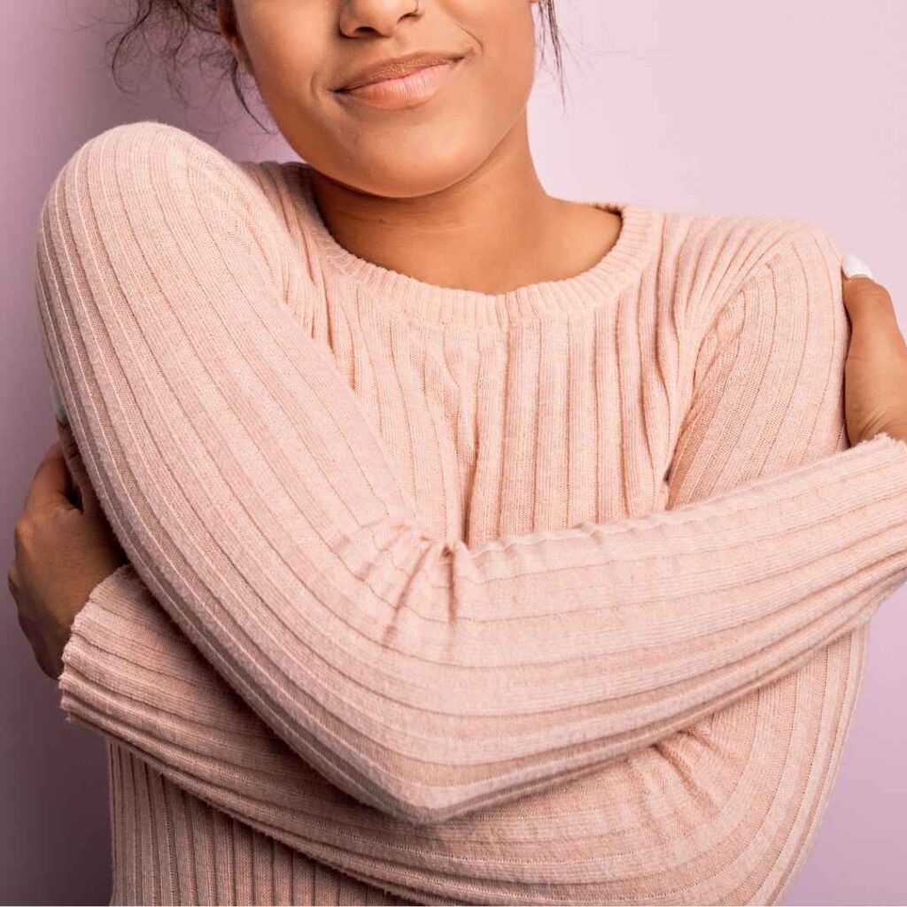 kvinde i lyserød sweater giver sig selv et kram som symbol på kropsbevidsthed, selvkærlighed og forbindelse til egen krop.