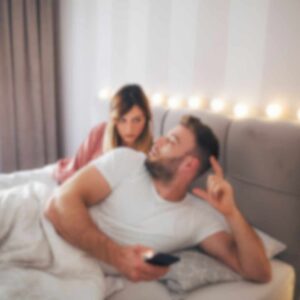 En kvinde kigger mistænksomt på sin partner, der ligger i sengen med sin telefon – et udtryk for jalousi i parforholdet og mistillid i relationen.