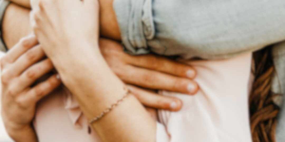 En mand omfavner en kvinde bagfra og holder hende tæt, hvilket kan symbolisere tryghed, kærlighed eller besiddende adfærd i et parforhold.